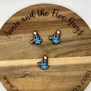 Three blue cartoon character pins with red hats and white wings placed on a round wooden board with text "Annie and the Five Guys" and a website link.