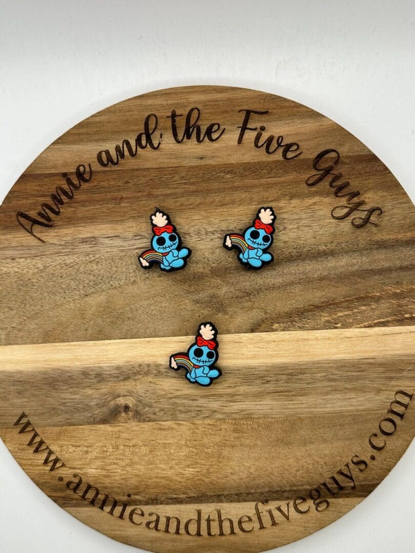 Three blue cartoon character pins with red hats and white wings placed on a round wooden board with text "Annie and the Five Guys" and a website link.