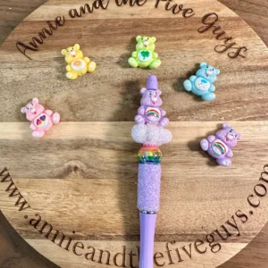 A purple pen with a glittery grip and rainbow decoration is placed on a round wooden board. Surrounding the purple pen are six colorful Caring Bears acrylic focal beads. The board’s text reads "Annie and the Five Guys.