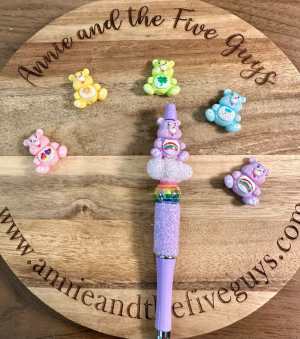 A purple pen with a glittery grip and rainbow decoration is placed on a round wooden board. Surrounding the purple pen are six colorful Caring Bears acrylic focal beads. The board’s text reads "Annie and the Five Guys.