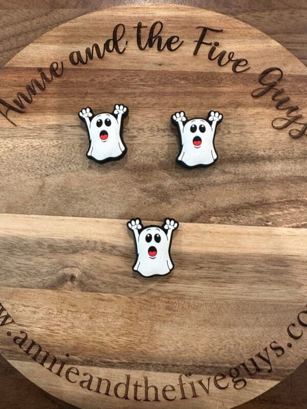 Three ghost-shaped cookies with raised arms, white frosting, and red mouths are arranged on a wooden board with the words "Annie and the Five Guys" engraved on it.