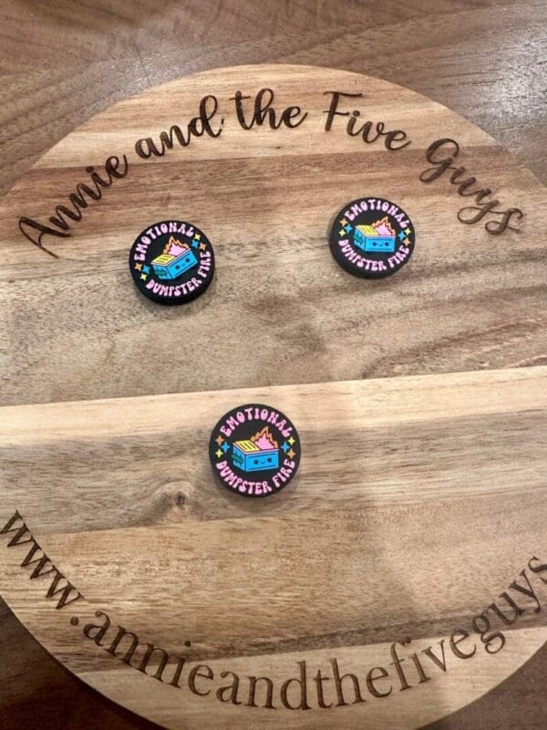 Three circular pins with "EMOTIONAL DUMPSTER FIRE" text and a graphic of a dumpster fire set on a wooden surface with "Annie and the Five Guys" text around the edge.

Replaced: Three Emotional Dumpster Fire silicone focal beads set on a wooden surface with "Annie and the Five Guys" text around the edge.