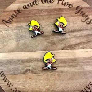 Three cartoon character pins with yellow hair and white outfits are displayed on a round wooden surface with the text "Annie and the Five Guys" and a web address.