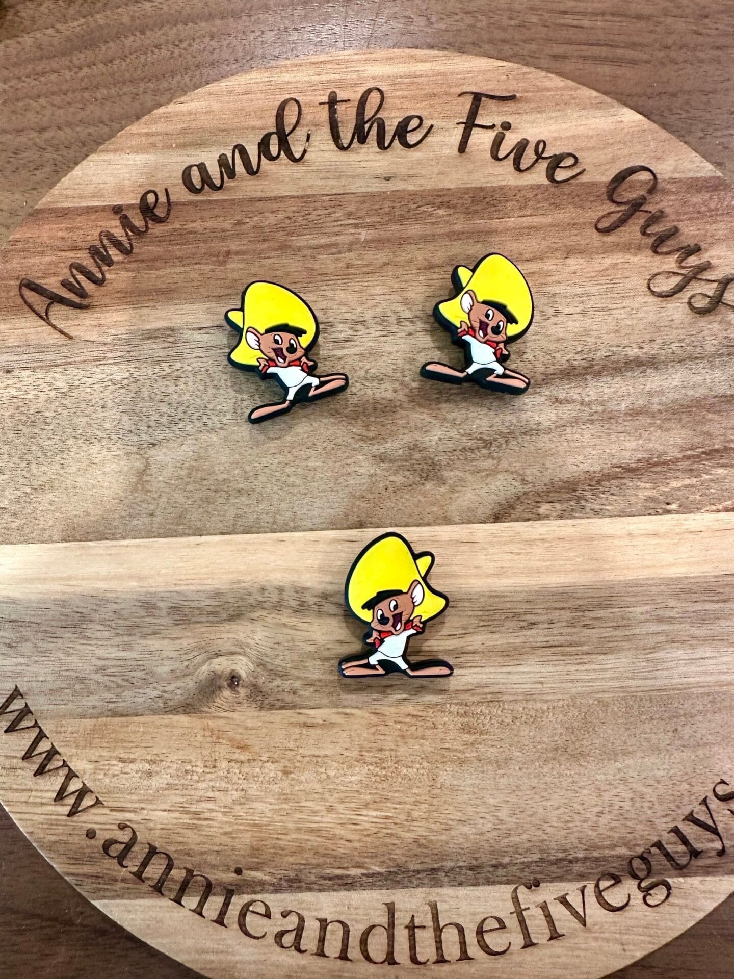 Three cartoon character pins with yellow hair and white outfits are displayed on a round wooden surface with the text "Annie and the Five Guys" and a web address.