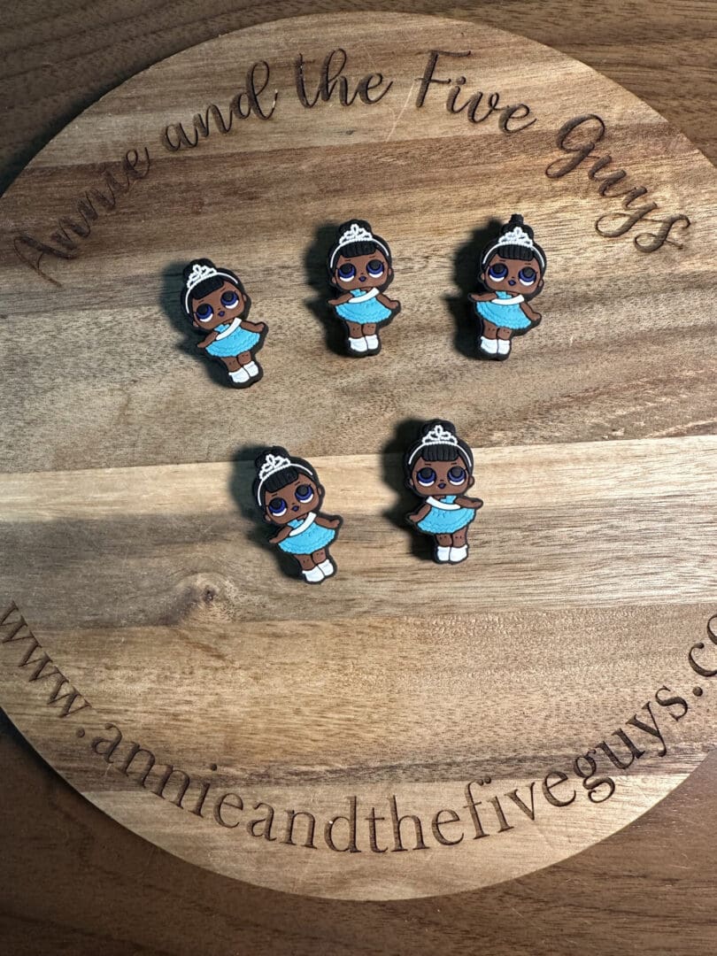 Four Princess T silicone focal beads of a girl with a tiara and blue dress on a circular wooden board with "Annie and the Five Guys" written around the edge.