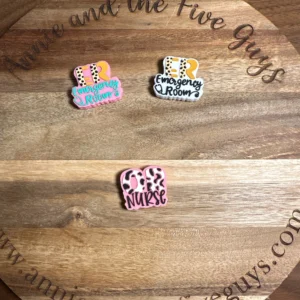 Three colorful Healthcare Silicone Focal Beads on a wooden surface. The beads read "ER Emergency Room" in different styles and colors, and "ER Nurse" in pink and white. The wood surface has engraved text around the edge.