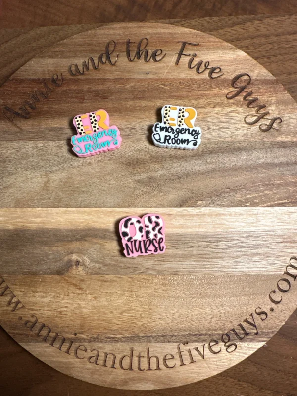 Three colorful Healthcare Silicone Focal Beads on a wooden surface. The beads read "ER Emergency Room" in different styles and colors, and "ER Nurse" in pink and white. The wood surface has engraved text around the edge.