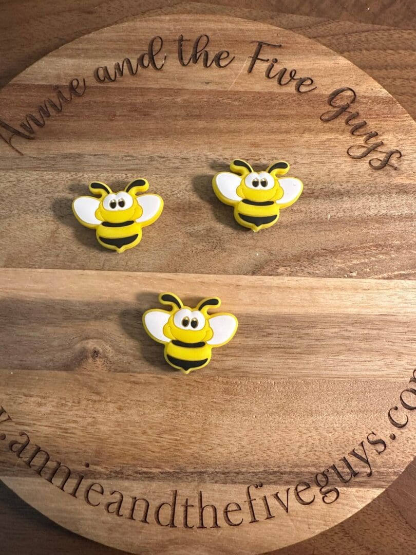 Three yellow and black bee-shaped magnets are arranged on a round wooden board with the text "Annie and the Five Guys" engraved around the edge.