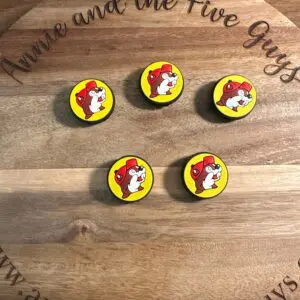 A wooden board with engraving "Buckeys silicone focal bead" displaying five circular pins featuring a cartoon dog wearing a red helmet. Website "www.annieandthefiveguys.com" is visible at the bottom.