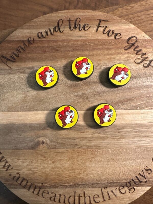 A wooden board with engraving "Buckeys silicone focal bead" displaying five circular pins featuring a cartoon dog wearing a red helmet. Website "www.annieandthefiveguys.com" is visible at the bottom.