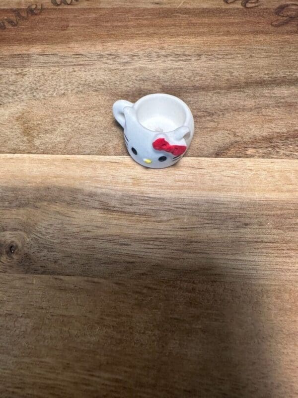 A small Sanrio cups resin bead shaped like a white cat with a red bow, placed on a wooden surface.