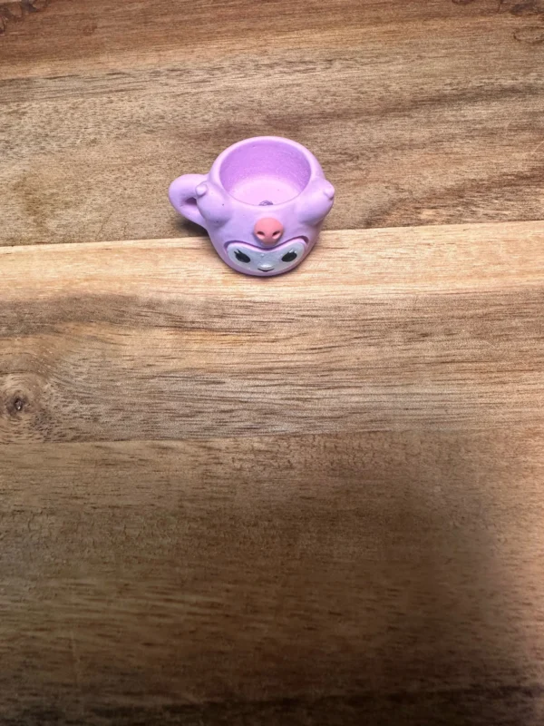 A small, purple Sanrio cups resin bead with eyes and small ears resembling an animal sits on a wooden surface.