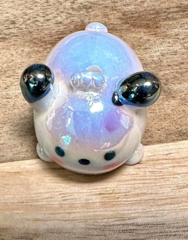 A small, shiny, Sanrio acrylic UV coated focal bead of a round, blue and white creature with black eyes and metallic silver ears, lying on a wooden surface.