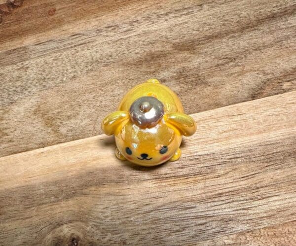 A small, yellow, Sanrio acrylic UV coated focal bead with a round head, large ears, and a shiny pearl-like decoration on its head, placed on a wooden surface.