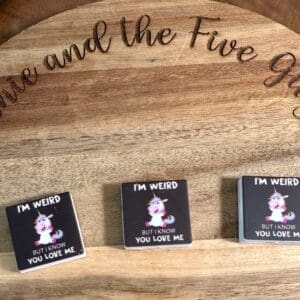 Three small square items are displayed on a wooden surface, each featuring the "I’m weird but I know you love me unicorn" illustration with text reading "I'm weird but I know you love me." The words "the Five" are partly visible in the background.