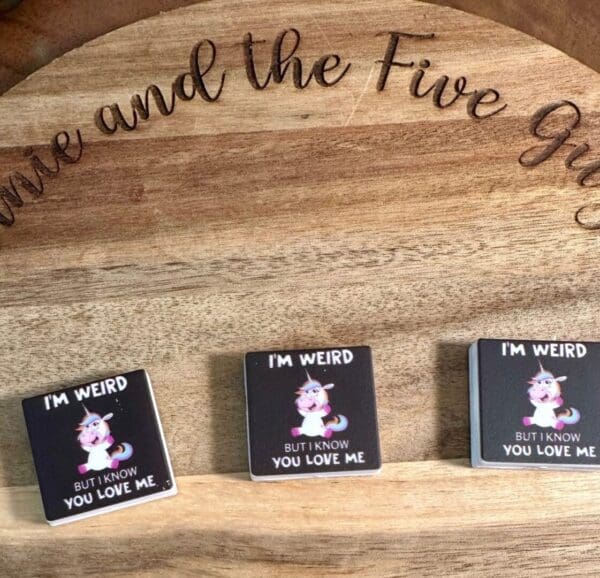 Three small square items are displayed on a wooden surface, each featuring the "I’m weird but I know you love me unicorn" illustration with text reading "I'm weird but I know you love me." The words "the Five" are partly visible in the background.