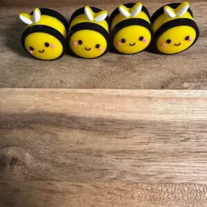 Four small, round, yellow 3D bee silicone focals with smiling faces and white wings are arranged in a line on a wooden surface.