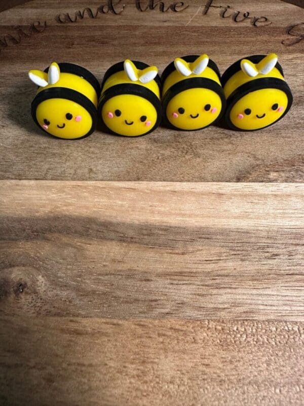 Four small, round, yellow 3D bee silicone focals with smiling faces and white wings are arranged in a line on a wooden surface.