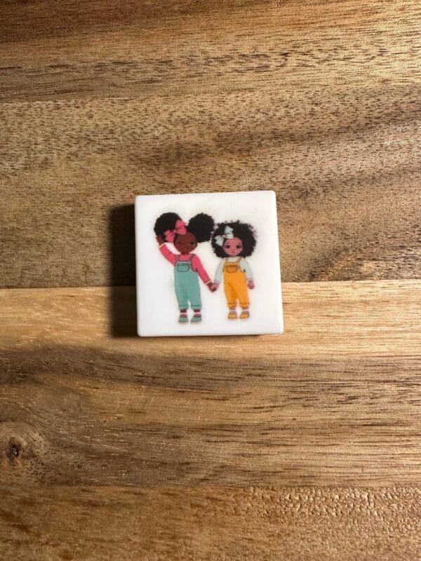 A small square tile with an illustration of two children holding hands, standing against a wooden background. One child is wearing a pink top with green pants, and the other is in a white top with yellow pants has been replaced by African American girl, women, Juneteenth silicone focal bead.