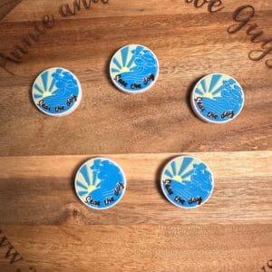 Five circular pins on a wooden surface with "annieandthefiveguys.com" written around the edge. The pins feature a blue bird with rays and the text "Seas the day".