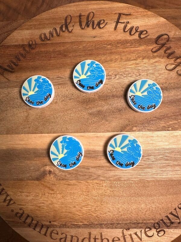 Five circular pins on a wooden surface with "annieandthefiveguys.com" written around the edge. The pins feature a blue bird with rays and the text "Seas the day".