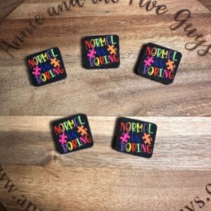 Five colorful pins with the phrase "Normal is Boring" in various sizes are displayed on a wooden surface with the text "annie and the Five Guys" visible.