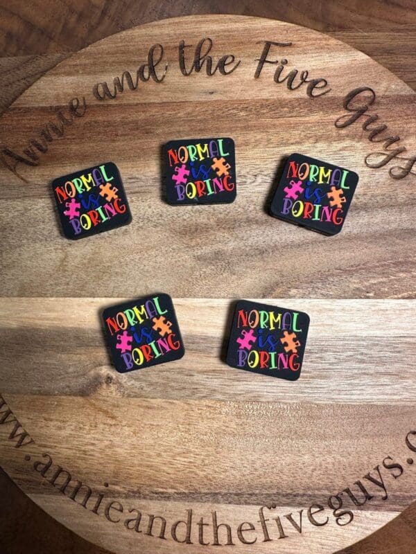 Five colorful pins with the phrase "Normal is Boring" in various sizes are displayed on a wooden surface with the text "annie and the Five Guys" visible.