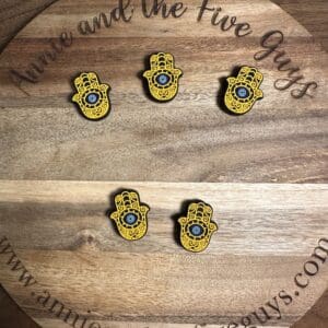 Five hamsa pins with blue eyes are arranged on a round wooden plaque engraved with the text "Annie and the Five Guys." The plaque is on a wooden surface.