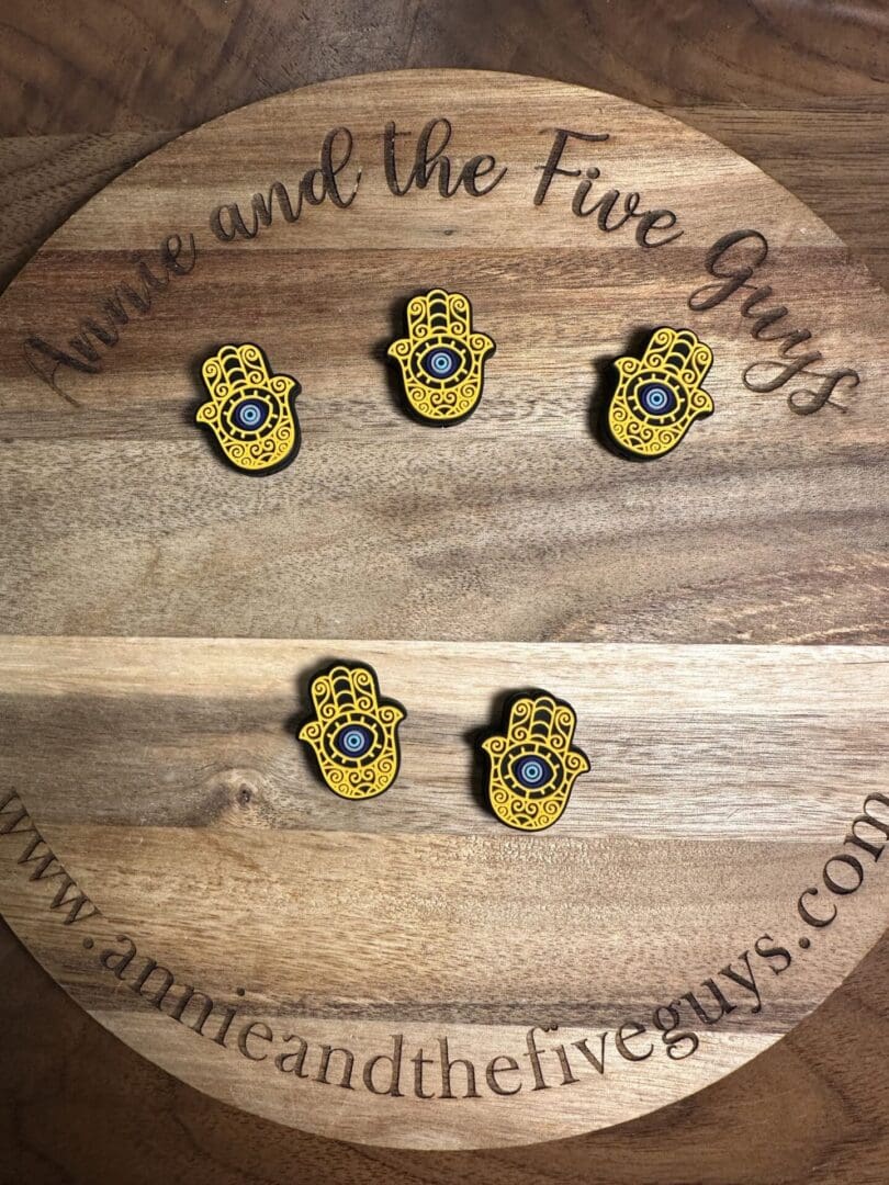 Five hamsa pins with blue eyes are arranged on a round wooden plaque engraved with the text "Annie and the Five Guys." The plaque is on a wooden surface.