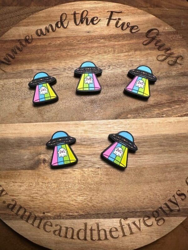 Five colorful Alien ghost ufo silicone focal beads featuring cartoon UFOs beaming up a ghost, arranged on a wooden board with text "annieandthefiveguys" at the bottom.