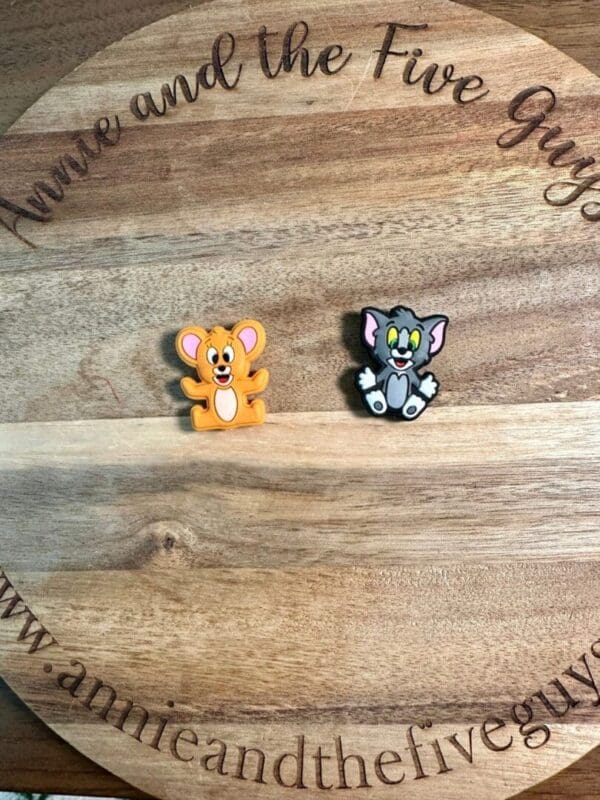 Two Cat and mouse silicone focal beads, one of a yellow mouse and the other a gray cat, placed on a circular wooden surface with text "Annie and the Five Guys" around the edge.