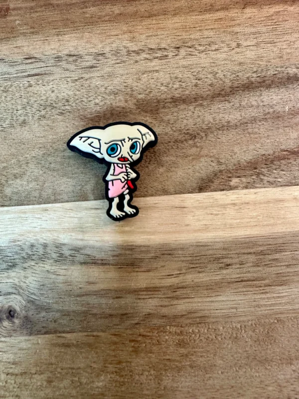 A small, cartoonish, humanoid figurine standing on a wooden surface. The figure has large ears, blue eyes, and is wearing a pink outfit, holding HP silicone focal beads.