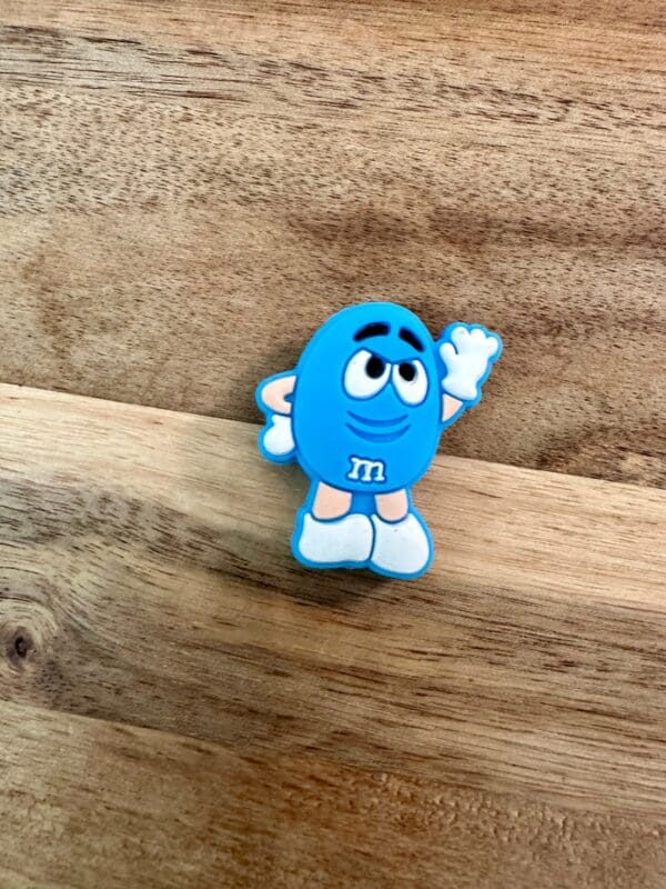 A small, blue Candy silicone focal bead with white gloves and shoes stands on a wooden surface, raising one hand.