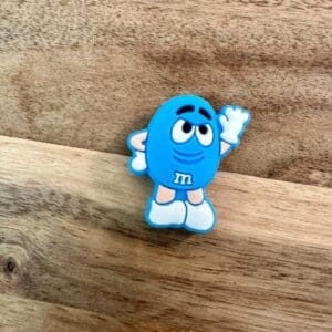 A small, blue Candy silicone focal bead with white gloves and shoes stands on a wooden surface, raising one hand.