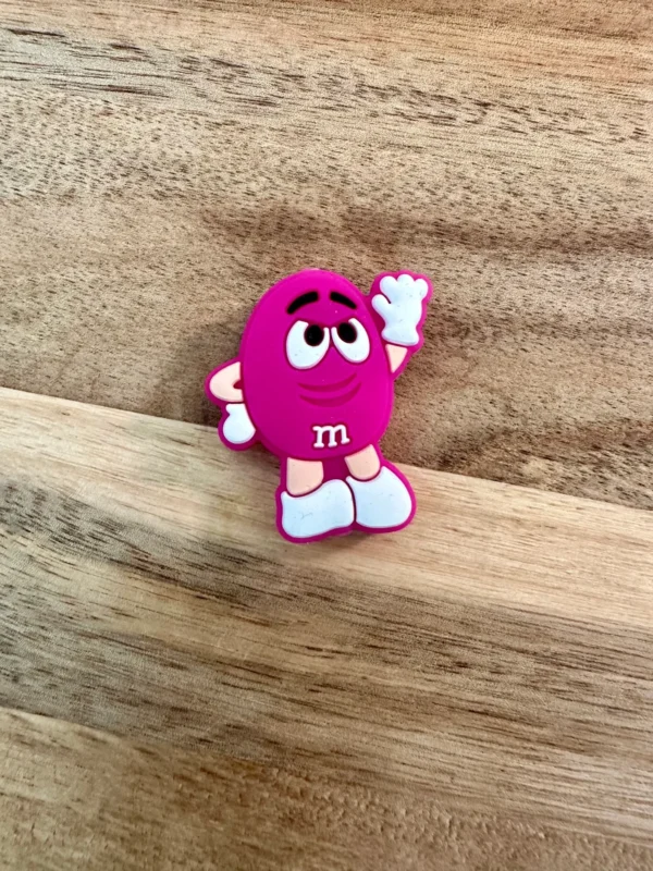 A purple Candy silicone focal bead with raised eyebrows, a white "m" on its front, and its right hand raised against a wooden surface.