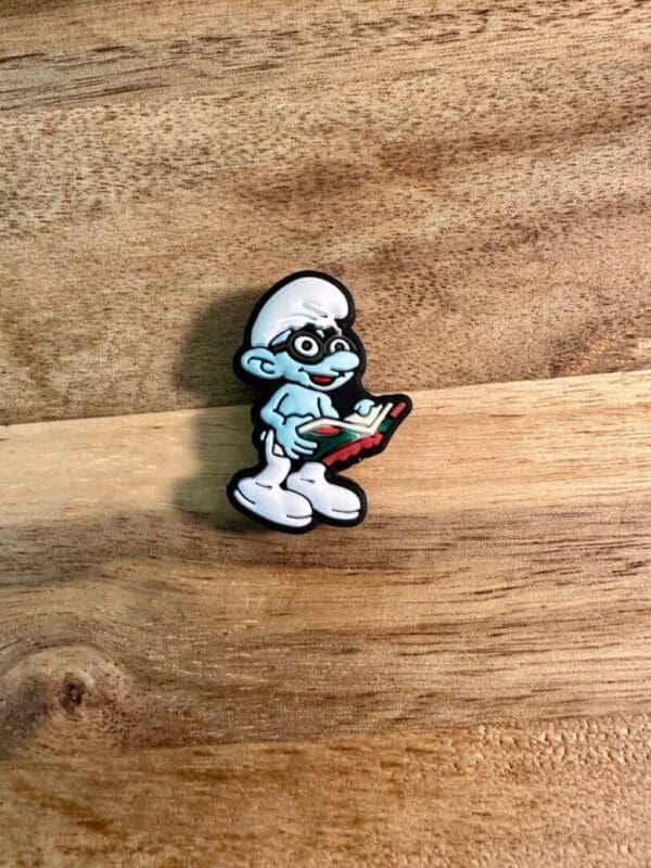 Blue people silicone focal bead of a Smurf wearing glasses and holding a book, placed on a wooden surface.