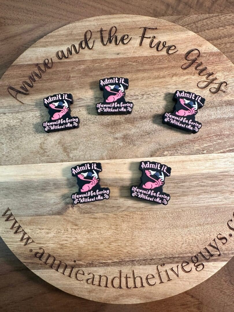 A wooden board with the text "Annie and the Five Guys" displays six Flamingo, funny silicone focal beads that read "Admit it. You would be boring without me." with a pink design. The website amyandthefiveguys.com is also visible.