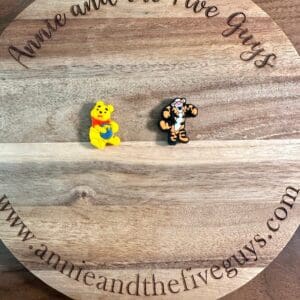 Two character figurines, one of a yellow bear and one of a tiger, are displayed on a circular wooden board with the text "Honey bear or tiger silicone focal bead" and a website URL.