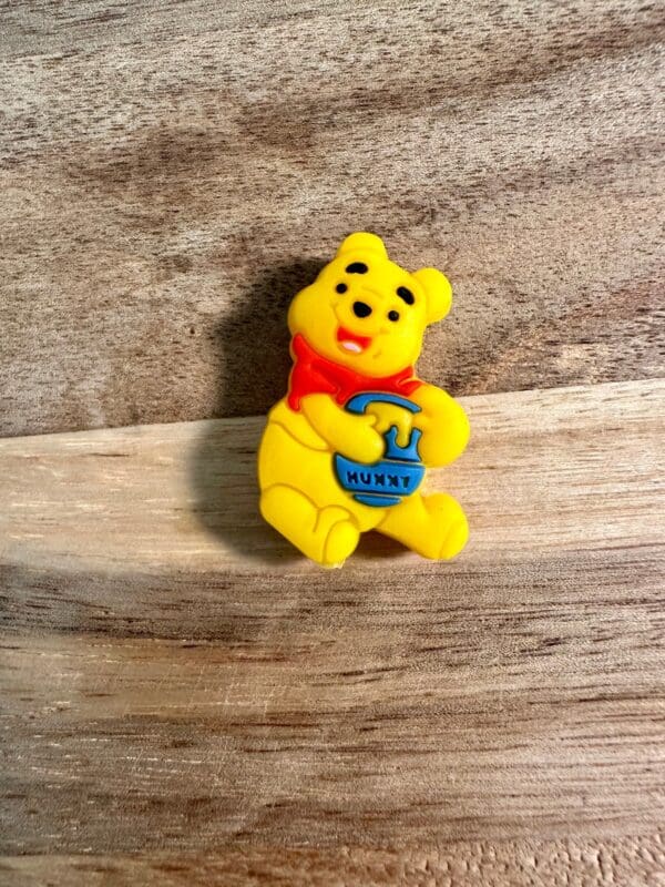 A small honey bear or tiger silicone focal bead, wearing a red shirt and holding a blue pot labeled "Hunny," sits on a wooden surface.