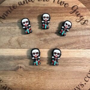 Five small *Saw silicone horror guy silicone focal beads* resembling the character from the horror movie "Saw" arranged on a wooden surface with text "Annie and the Five Guys" engraved on it.