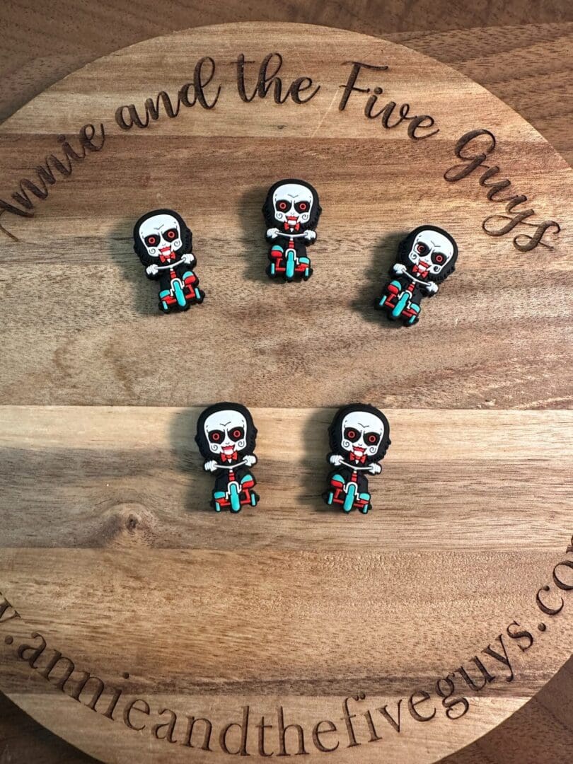 Five small *Saw silicone horror guy silicone focal beads* resembling the character from the horror movie "Saw" arranged on a wooden surface with text "Annie and the Five Guys" engraved on it.