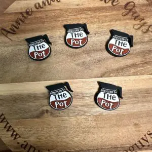 Five "Addicted to the Pot" pins with a cooking pot design displayed on a wooden board engraved with "Annie and the Five Guys" and the website URL "www.annieandthefiveguys.com".