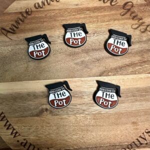 Five "Addicted to the Pot" pins with a cooking pot design displayed on a wooden board engraved with "Annie and the Five Guys" and the website URL "www.annieandthefiveguys.com".