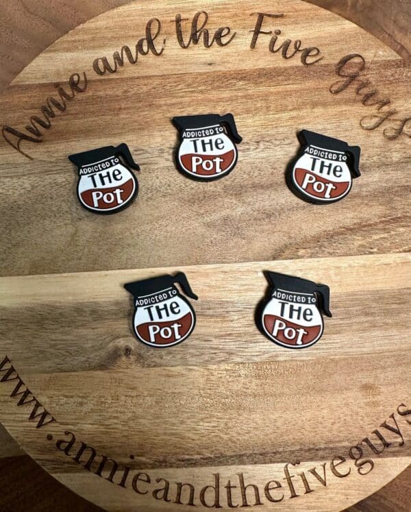 Five "Addicted to the Pot" pins with a cooking pot design displayed on a wooden board engraved with "Annie and the Five Guys" and the website URL "www.annieandthefiveguys.com".