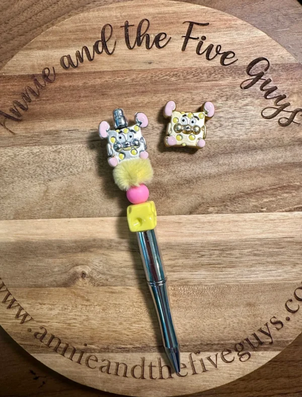 A Mouse cheese bead with a fluffy, decorated topper resembling a mouse and a square-shaped mouse-themed charm placed on a wooden surface with the engraved text "Annie and the Five Guys".