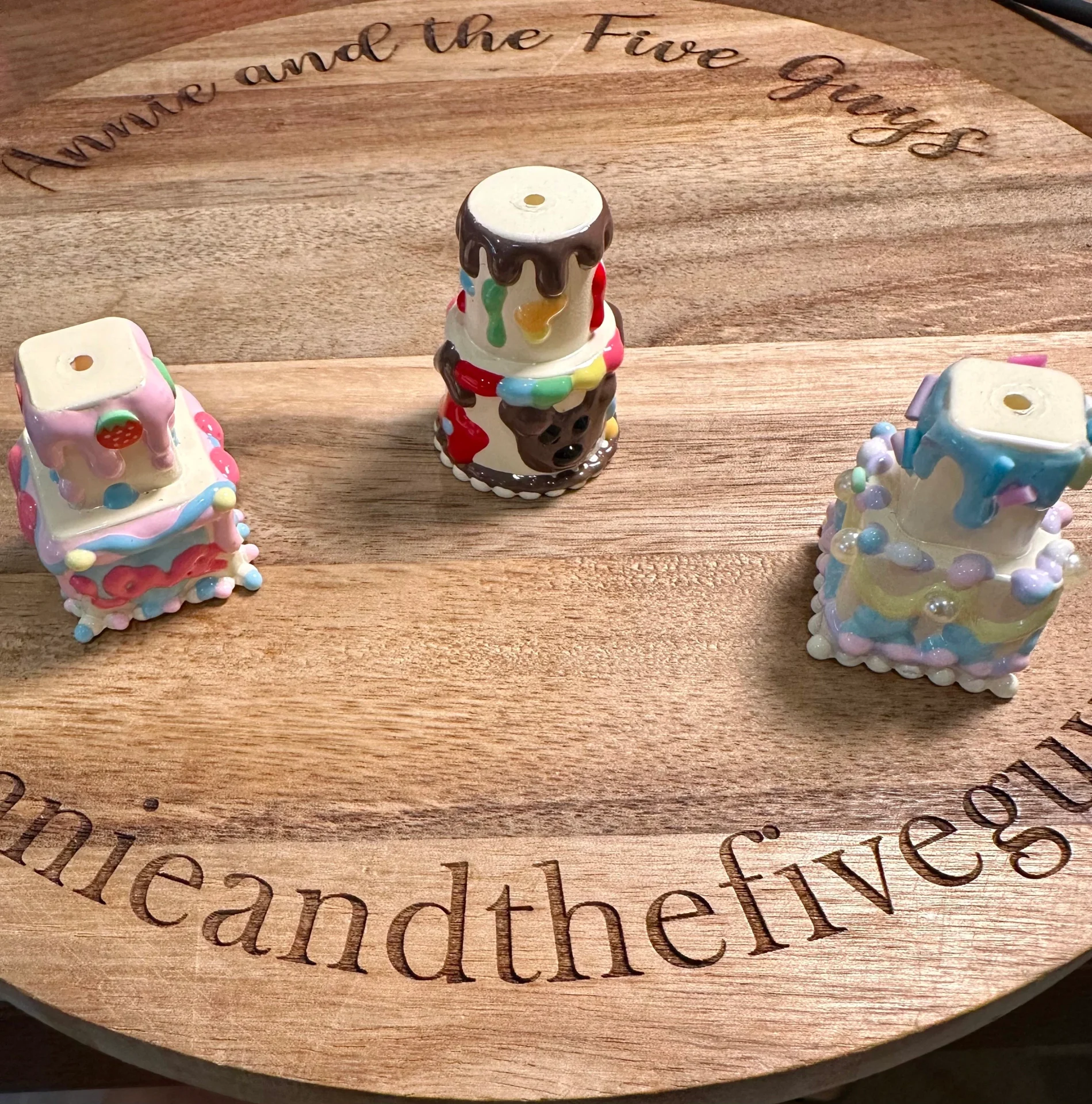 Three 3d cake focal beads with colorful icing and decorations are displayed on a wooden surface inscribed with "Annie and the Five Guys.