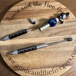 A wooden round tray with the text "Annie and the Five Guys" holds a beadable pen, stylus, ruler, screwdriver in one, a ballpoint pen in a plastic cover, and a small figurine seated next to a sphere.