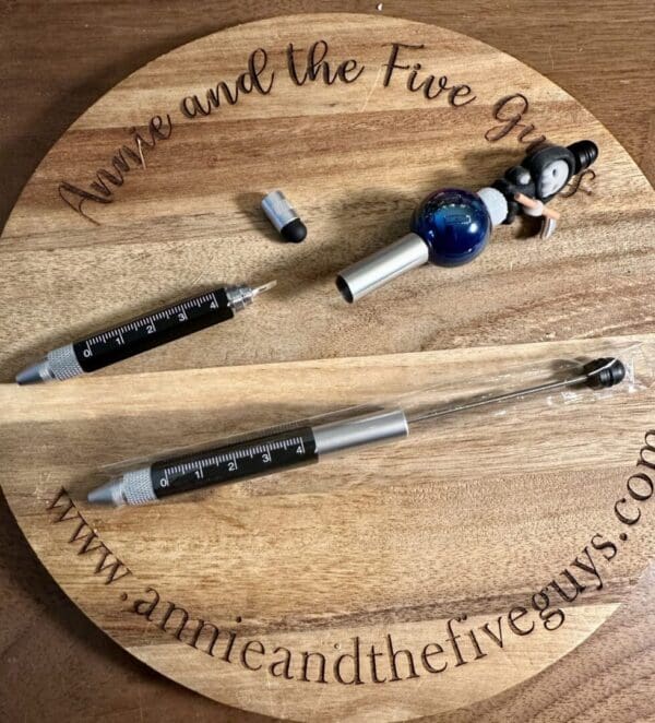 A wooden round tray with the text "Annie and the Five Guys" holds a beadable pen, stylus, ruler, screwdriver in one, a ballpoint pen in a plastic cover, and a small figurine seated next to a sphere.