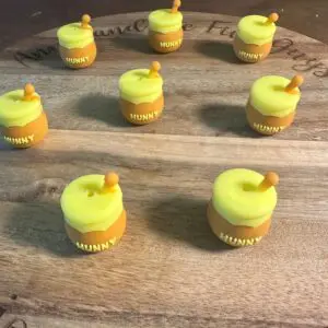 Seven small candles shaped like honey pot silicone focal beads labeled "Hunny" are arranged in a circular pattern on a wooden board.