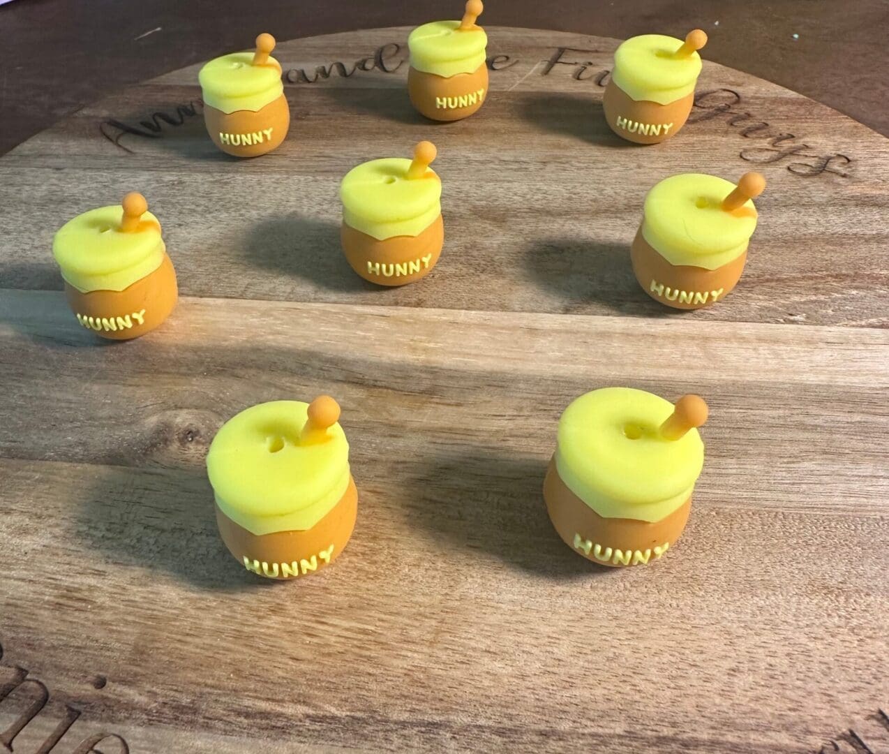 Seven small candles shaped like honey pot silicone focal beads labeled "Hunny" are arranged in a circular pattern on a wooden board.